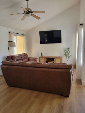 Ein Wohnzimmer mit braunem Ledersofa, Deckenventilator, Flachbildfernseher, Stehlampe und einer kleinen Pflanze.