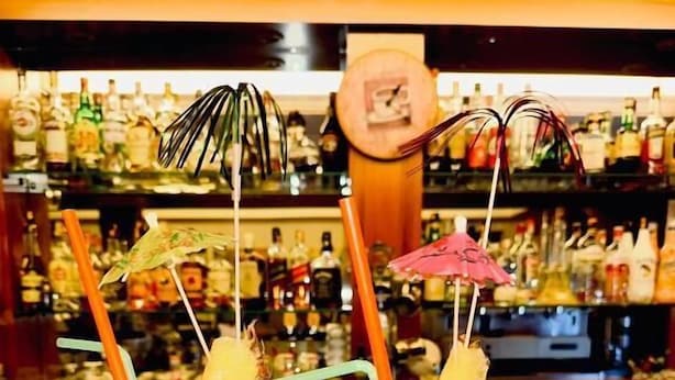 Zwei Cocktails mit Trinkhalmen und Schirmchen auf einer Bar, im Hintergrund verschiedene Flaschen.