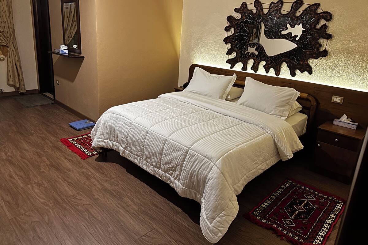 A neatly made bed with a white comforter and pillows, a wooden headboard, a bedside table with a tissue box, a red patterned rug, and a wall-mounted decorative piece.