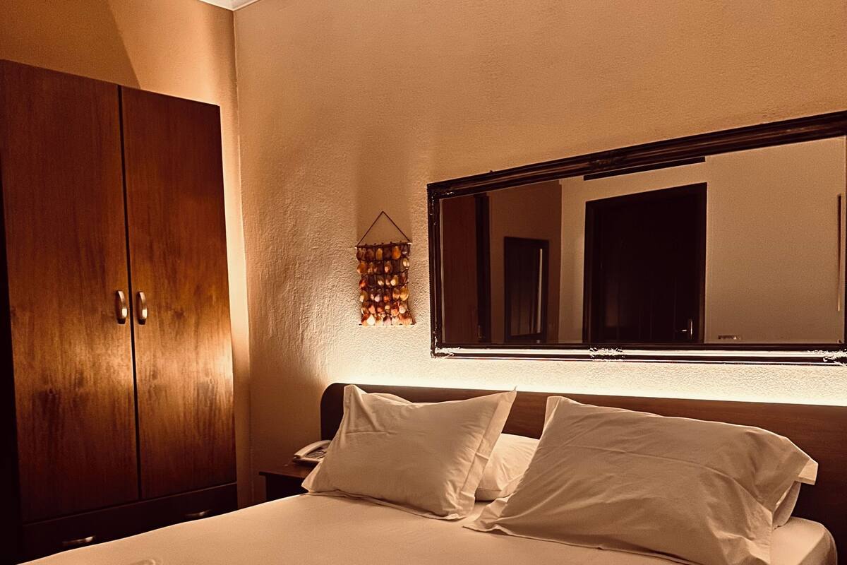 A neatly made bed with pillows, a wooden wardrobe, and a wall-mounted mirror.