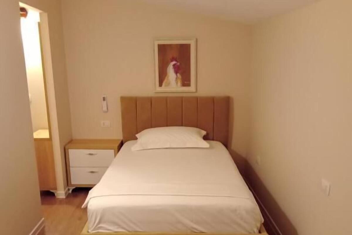 A bedroom with a bed, a nightstand, a wall-mounted light switch, and a framed picture above the bed.