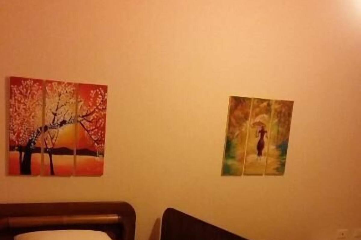 A hotel room with two single beds, a wooden headboard, and a painting of a tree on the wall.