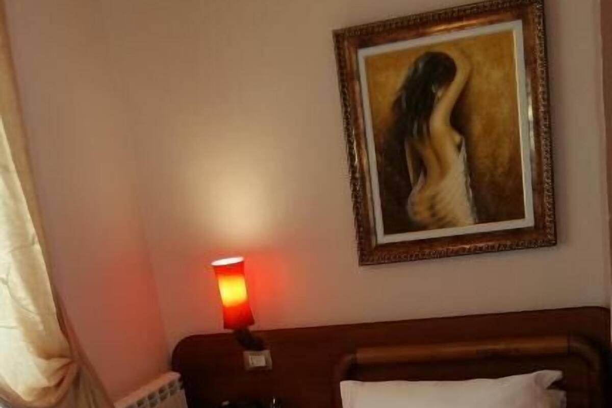 A single bed with white linens, a nightstand with a lamp, a framed painting on the wall, and a radiator.