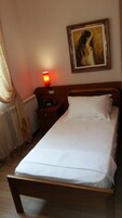 Una cama individual con sábanas blancas, una mesita de noche con lámpara, un cuadro en la pared y un radiador.
