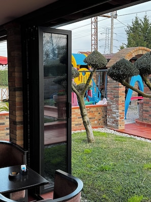 A modern outdoor seating area with a glass partition, a brick pillar, and a view of a children's play area with colorful structures.
