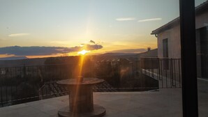 Un balcon avec une balustrade, une table et une vue sur un coucher de soleil au-dessus d’une étendue d’eau et de collines lointaines.