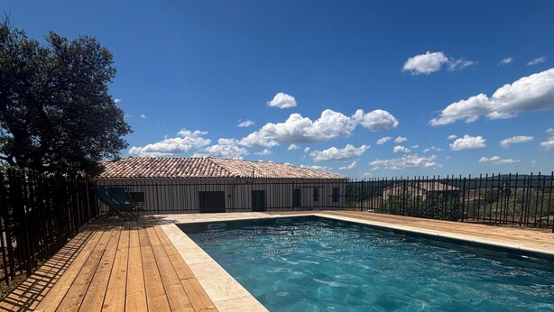 Une terrasse en bois avec une piscine, entourée d’une clôture et d’un bâtiment dont le toit est recouvert de tuiles.