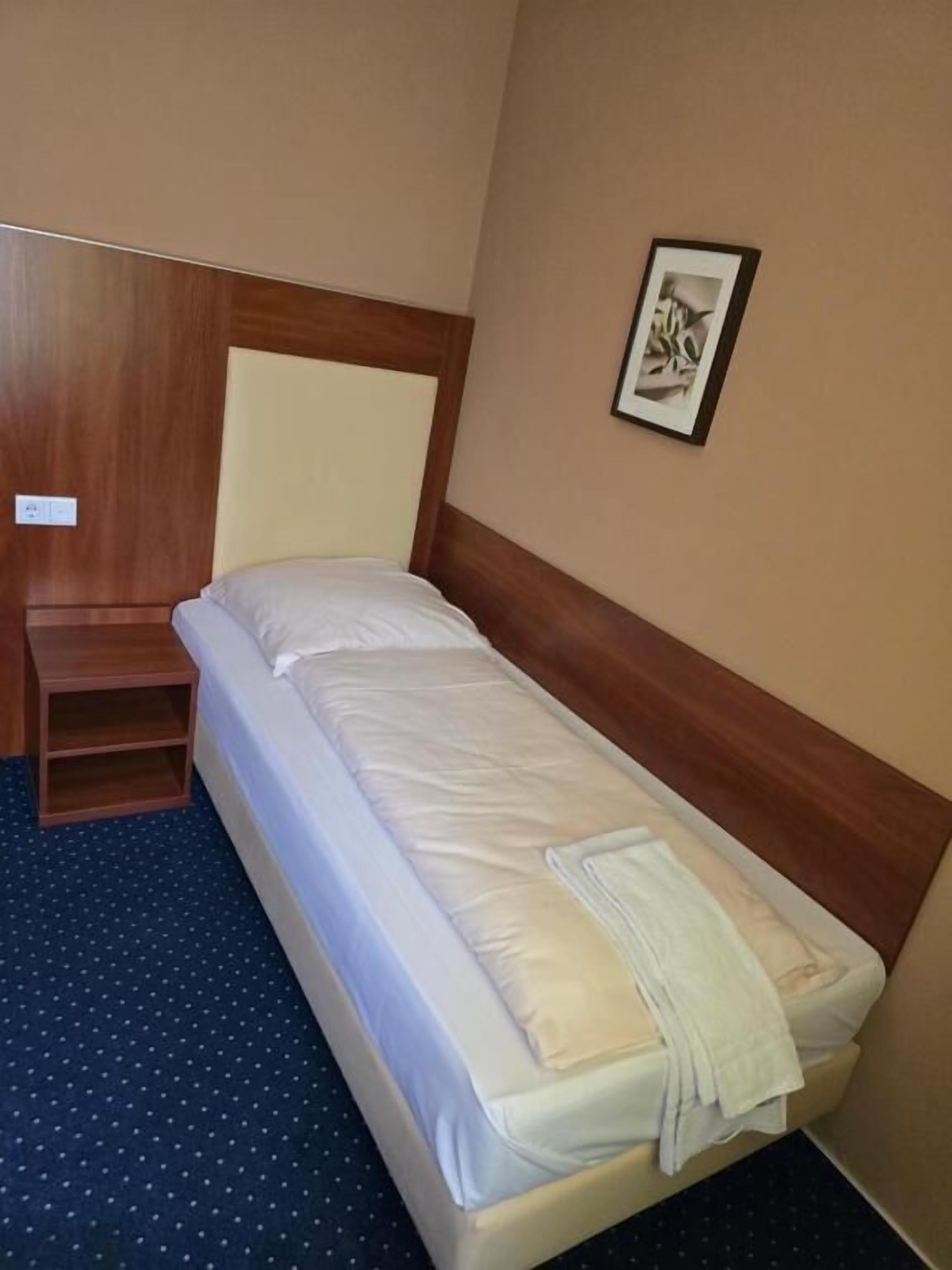 Ein Einzelbett mit Holz-Kopfteil, ein kleiner Nachttisch aus Holz und ein gerahmtes Bild an der Wand.