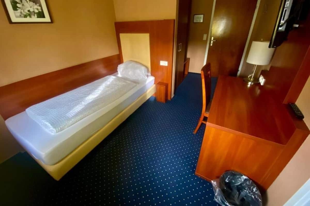 A hotel room with a single bed, a desk, a chair, a trash bin, and a door.