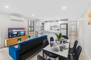 Una cocina moderna con armarios blancos, un área de comedor con un sofá azul y un televisor que muestra varios canales.