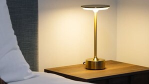 Un lit bien fait, avec du linge de lit blanc, une table de chevet en bois surmontée d’une lampe moderne et une tête de lit grise.