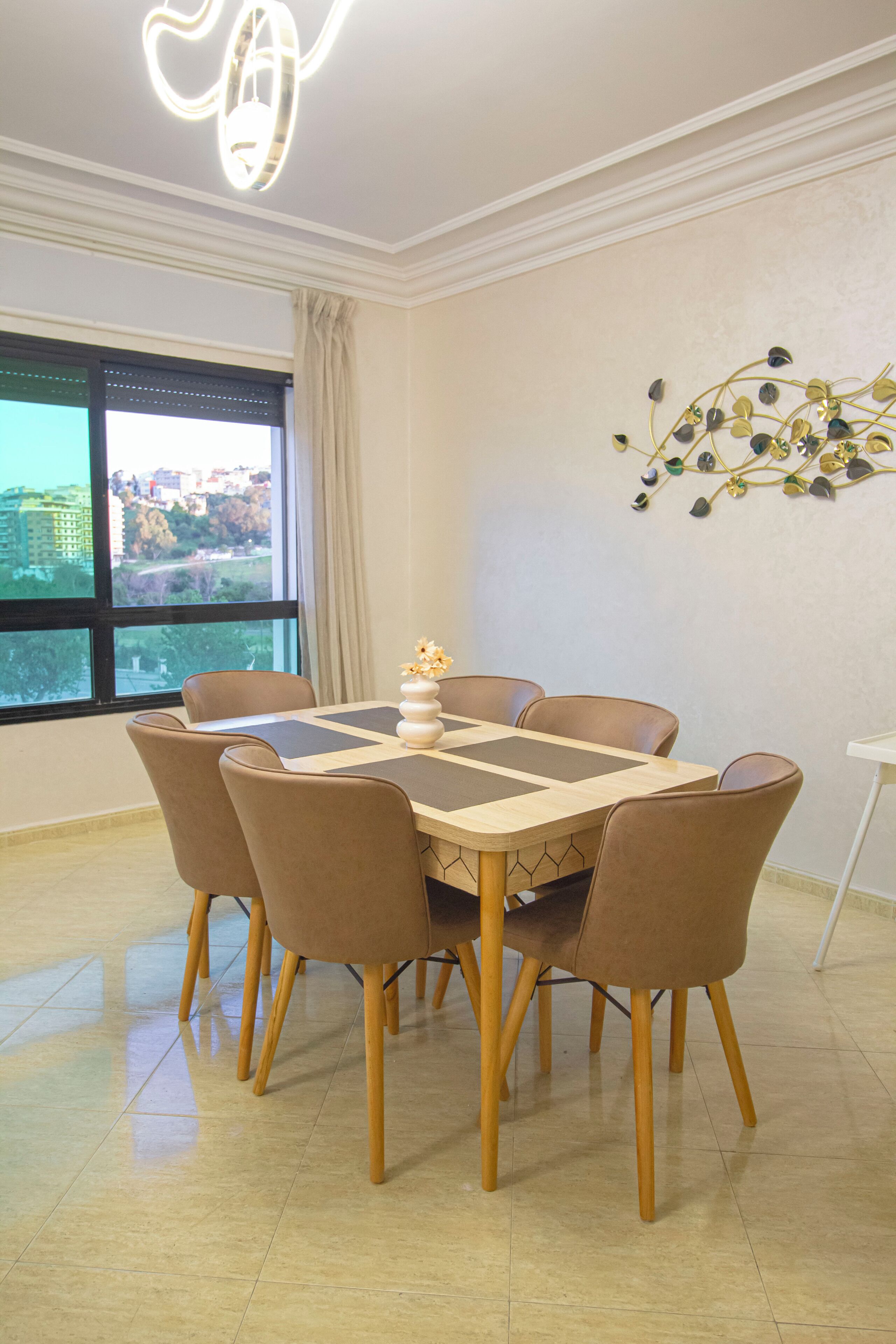 A dining area with a table and chairs, a chandelier, a window with a view, and a wall decoration.