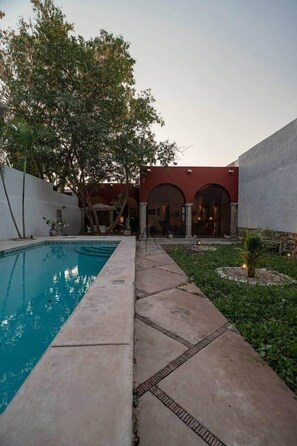 A swimming pool with a paved walkway, a building with arched entrances, and a tree in the background. - Casa Luz: Cozy Retreat + Pool (Mérida)