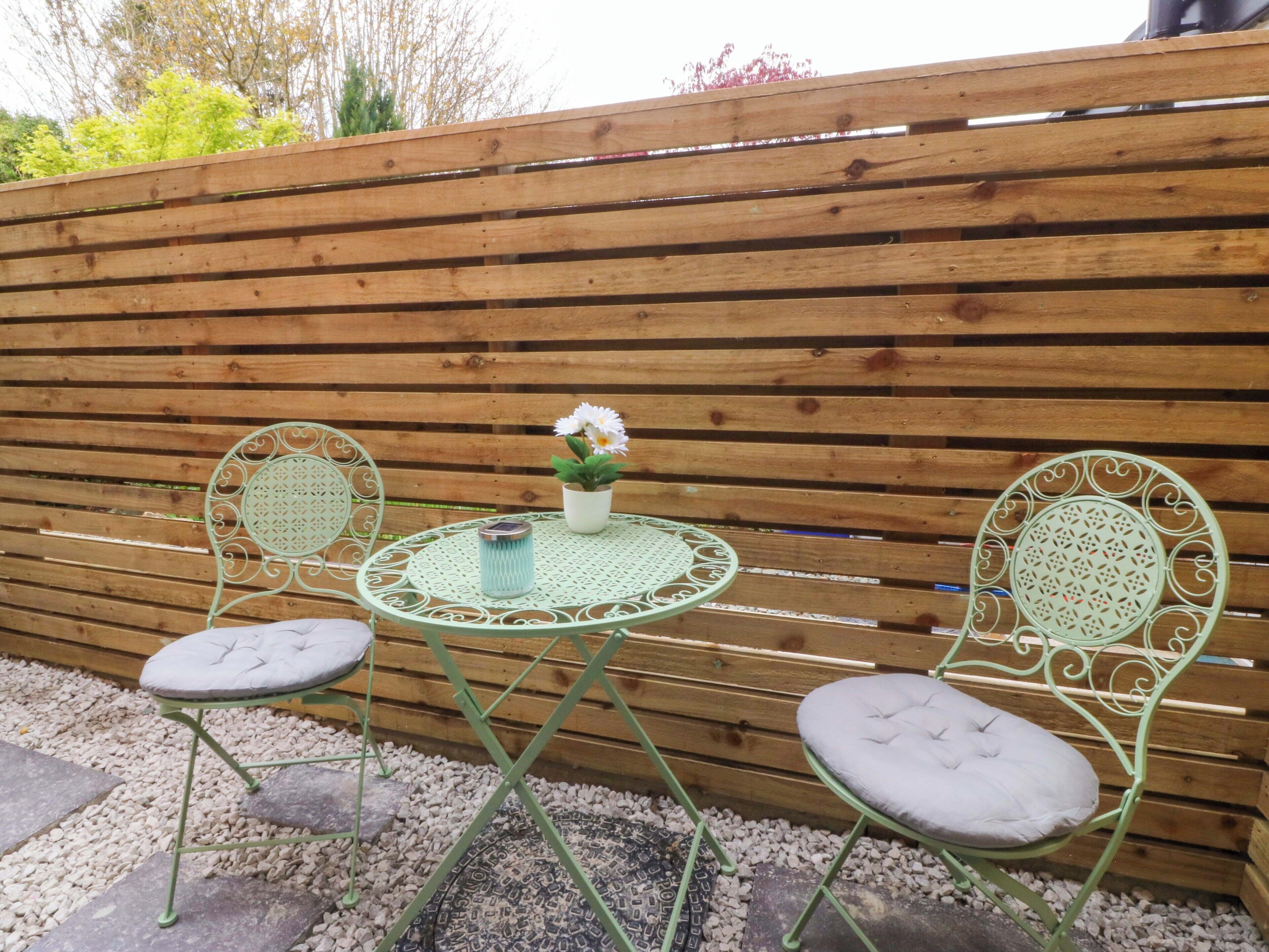 A small outdoor seating area with a round table and two chairs, a potted plant, and a wooden fence in the background.