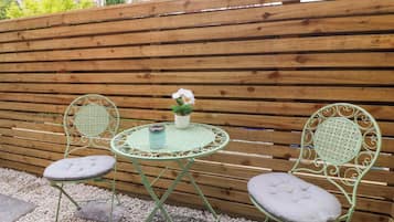 A small outdoor seating area with a round table and two chairs, a potted plant, and a wooden fence in the background.