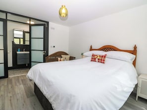 A bedroom with a large bed, a wooden headboard, a nightstand, a bedside lamp, a mirror, and a bathroom with a sink and toilet. - Trwyn Cam (Caernarfon)