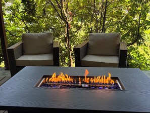 A rectangular outdoor fire feature with flames, flanked by two cushioned chairs with a wicker frame, set against a backdrop of dense green foliage. - SoHo-Lakeside Retreat (Abingdon)