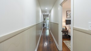 A narrow hallway with wooden flooring, white walls, and a door leading to a bedroom with a bed, nightstand, and lamp. - Murfreesboro Gem Near Square + MTSU - Cozy Cottage (Murfreesboro)