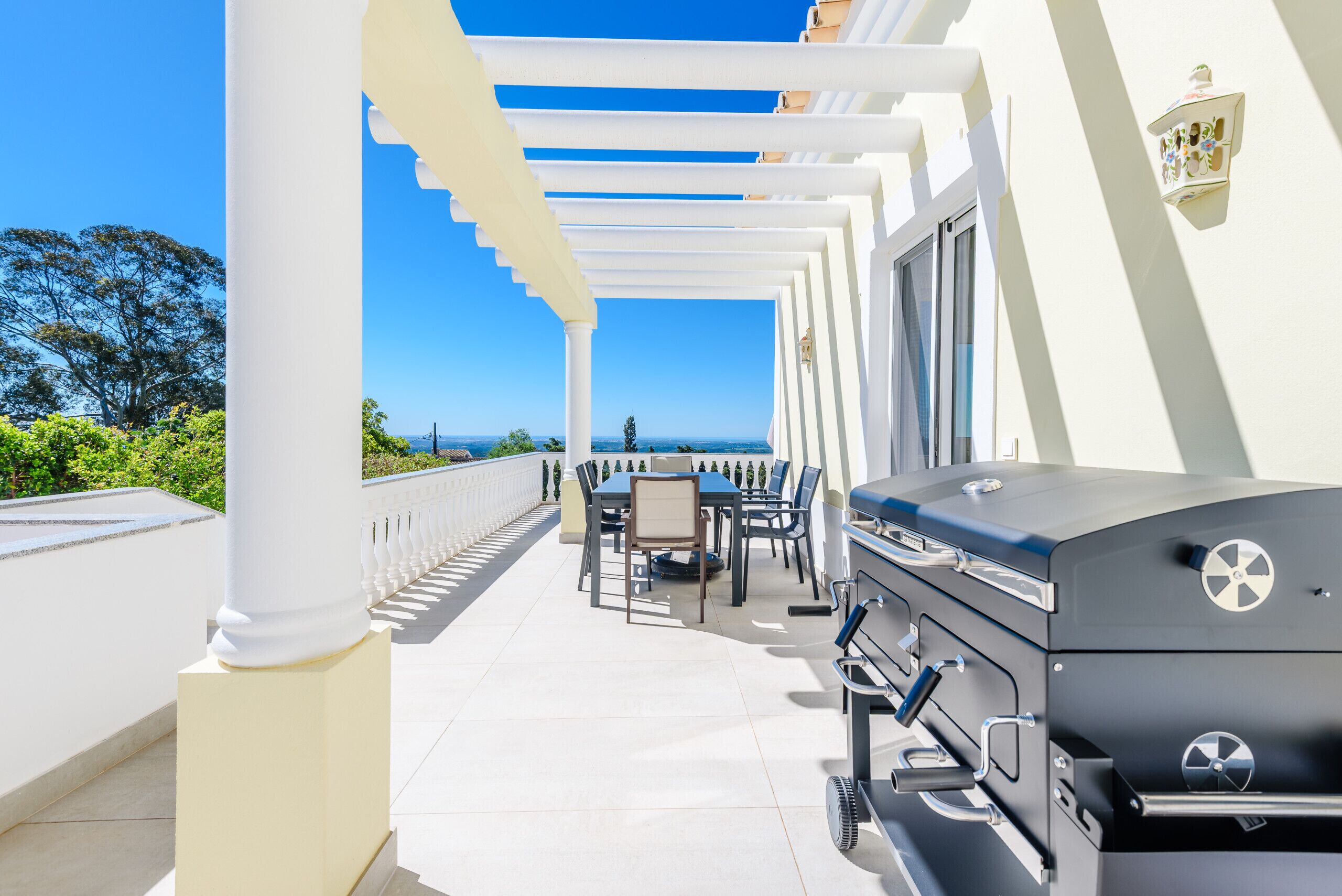 A covered outdoor patio with a grill, dining table, and chairs, overlooking a scenic view.