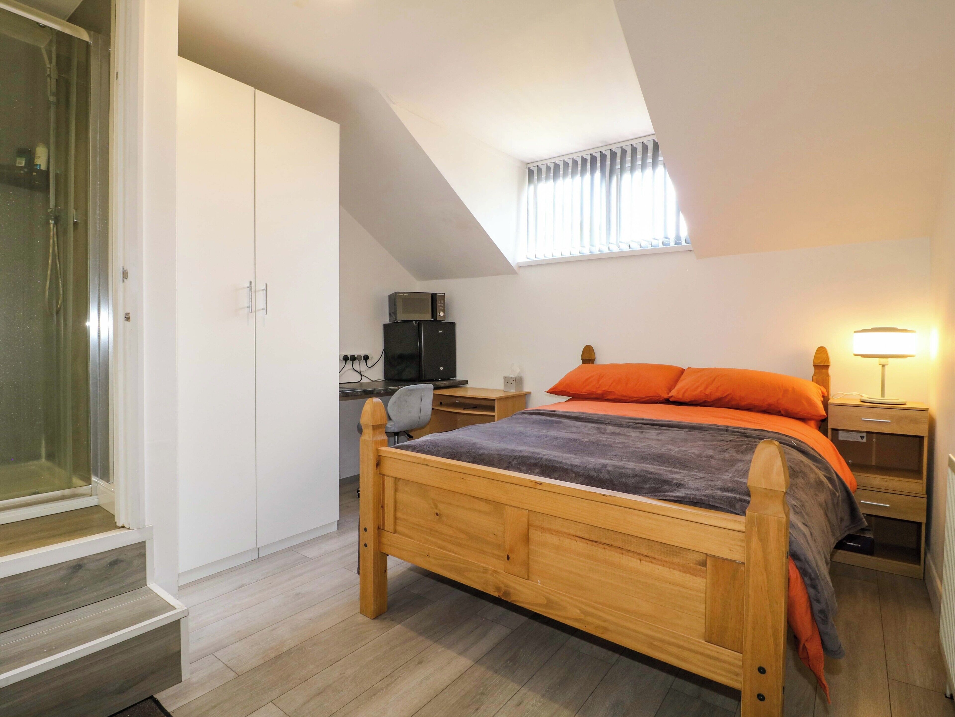Une chambre avec un lit en bois, un bureau avec un ordinateur, un four à micro-ondes et une fenêtre avec des rideaux.