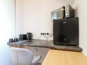 Una kitchenette con microondas, hervidor de agua y cafetera sobre una encimera de mármol, una silla y un enchufe.