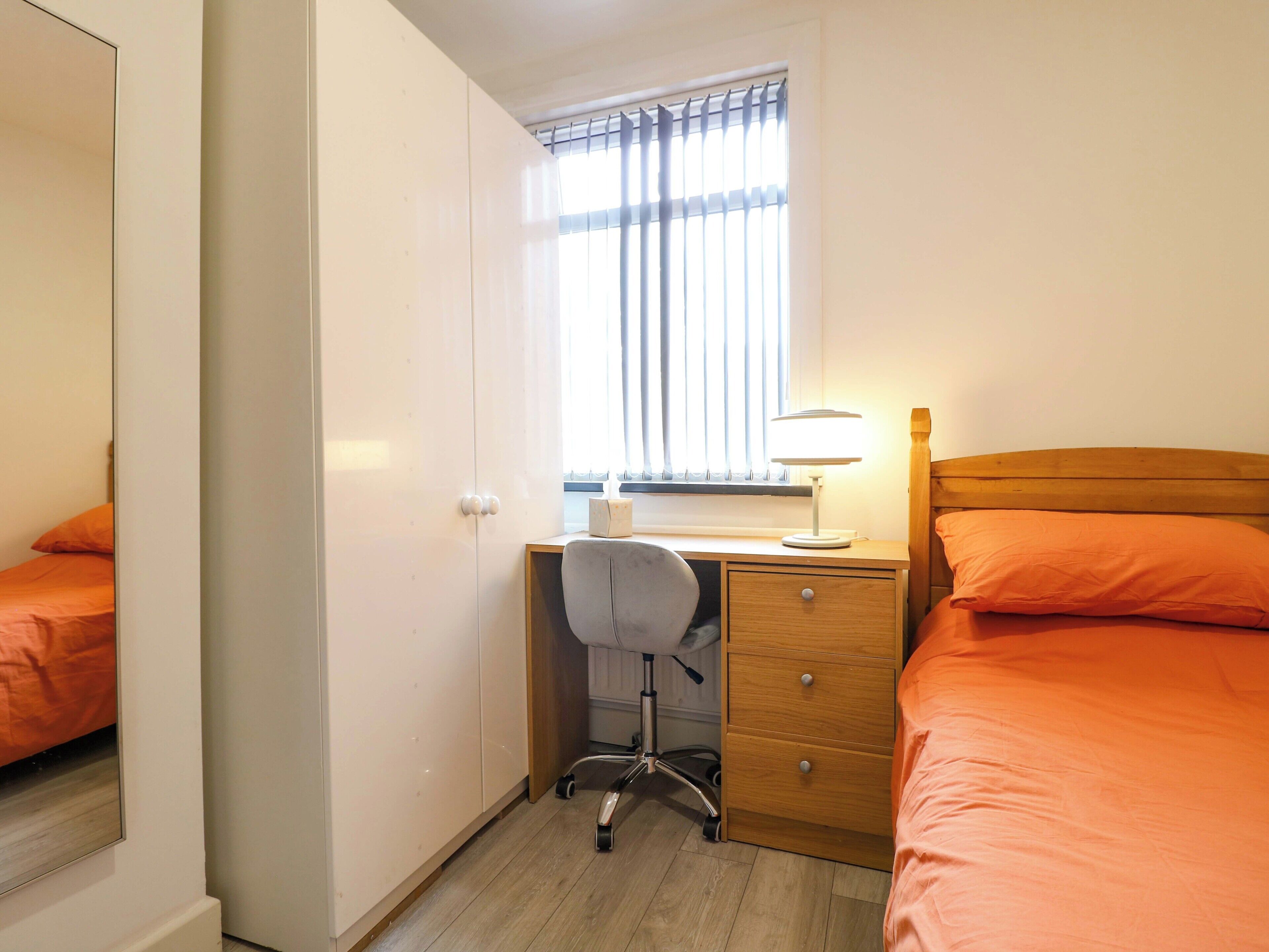 A bedroom with a bed, a desk with a chair, a nightstand, and a window with blinds.
