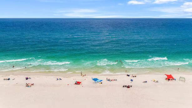 Una playa con aguas azul cristalino, gente tomando el sol y sombrillas.