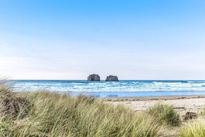 Une plage avec deux formations rocheuses bien visibles au loin, un ciel dégagé et un premier plan sablonneux parsemé d’herbe.