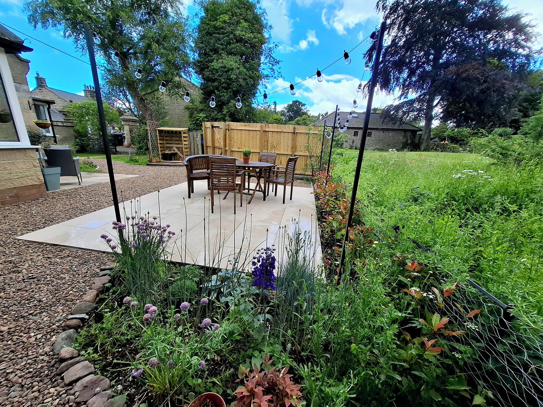 A patio with a table and chairs, surrounded by a garden with various plants and a wooden fence.