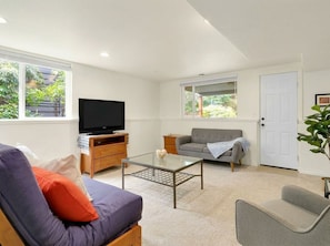 A living room with a sofa, armchair, glass coffee table, television, and a door. - 2-bedroom apartment in peaceful Seattle neighborhood near beach and downtown (Seattle)