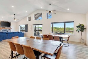 A spacious room with a large dining table, comfortable seating, a TV, and a view of the outdoors. - SDC | Lake View | Pools·Hot Tub·Sauna | Theater Rm (MARVEL CAV PK)