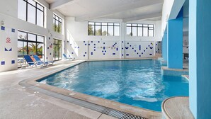 Una piscina interior con agua azul cristalina, rodeada de paredes de azulejos azules y blancos, y equipada con tumbonas.