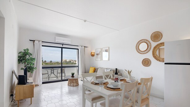 Un comedor moderno con una mesa blanca dispuesta para dos comensales, un sofá y un balcón con vistas.
