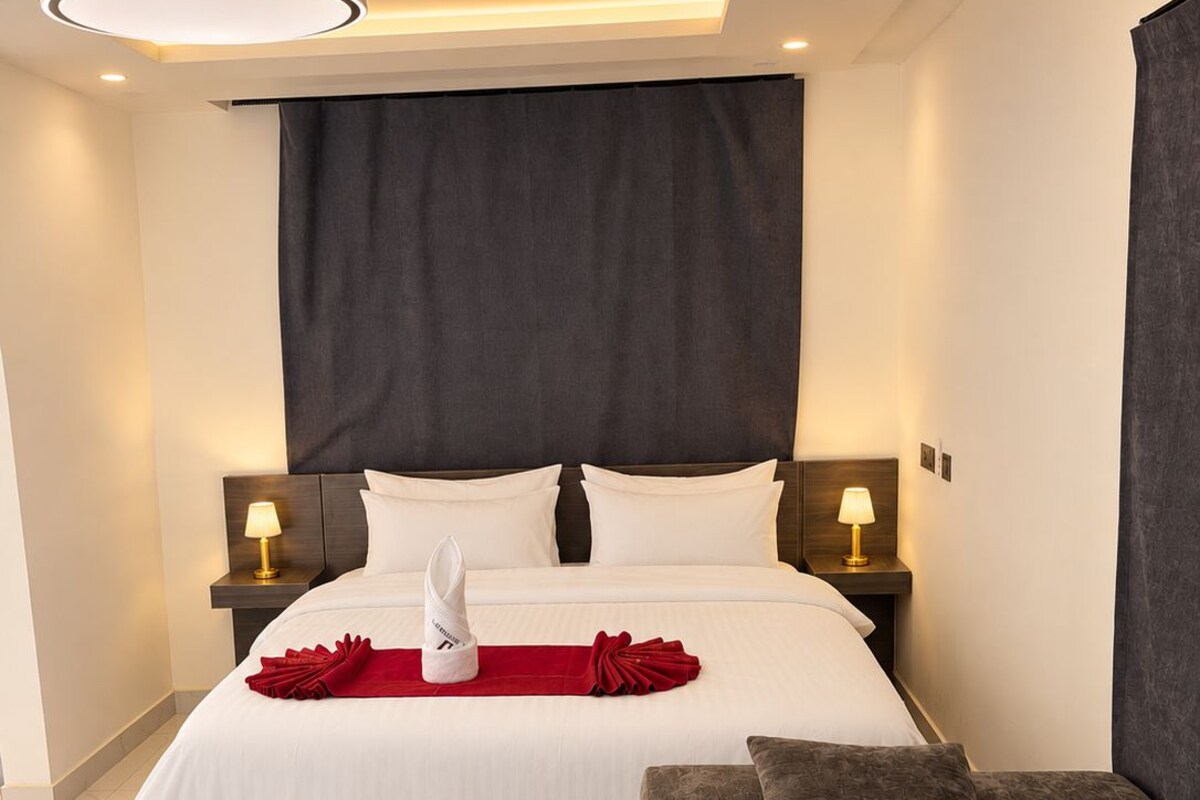 A modern hotel room with a large bed, two bedside tables with lamps, a dark headboard, a red towel, a grey sofa, and a blue patterned rug.