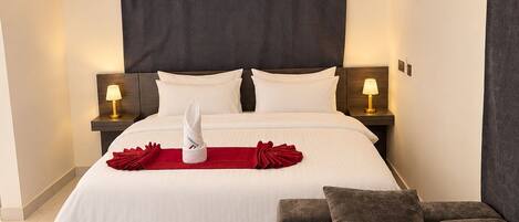 A modern hotel room with a large bed, two bedside tables with lamps, a dark headboard, a red towel, a grey sofa, and a blue patterned rug.