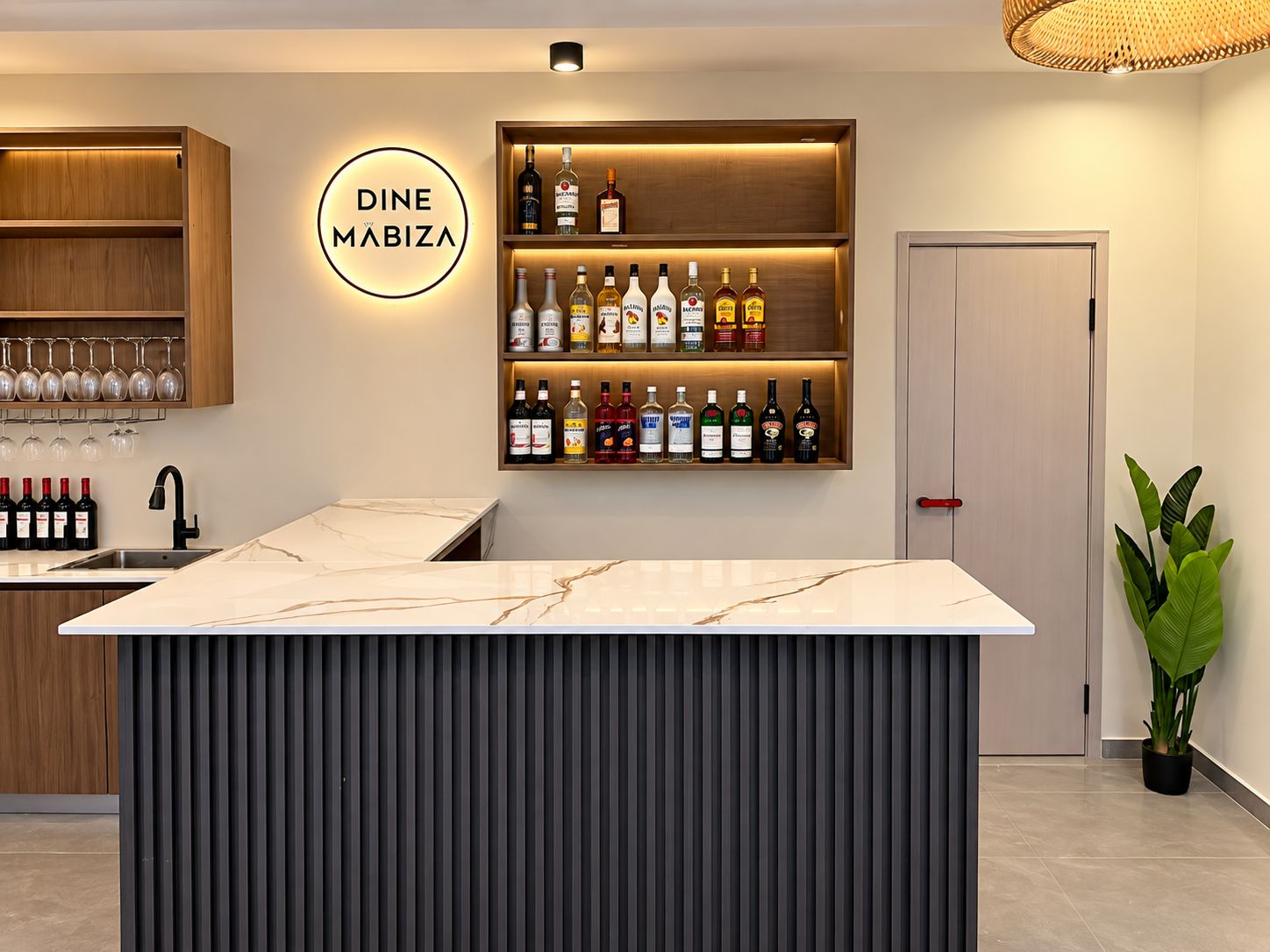 Un espace bar doté d’un comptoir en marbre, d’étagères remplies de bouteilles d’alcool et d’une enseigne indiquant : « DINE MABIZA ».
