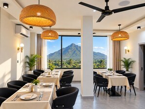 Un comedor con mesas dispuestas para una comida, ventanas amplias con vista a la montaña y lámparas de mimbre colgantes.