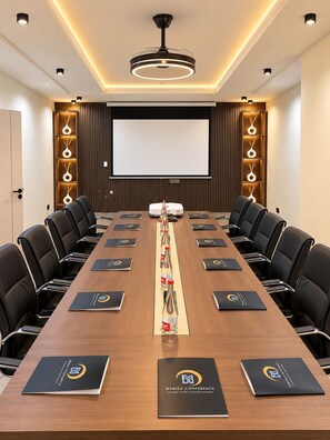 Un salón de conferencias con una larga mesa, sillas negras, una pantalla de proyector y una lámpara en el techo.