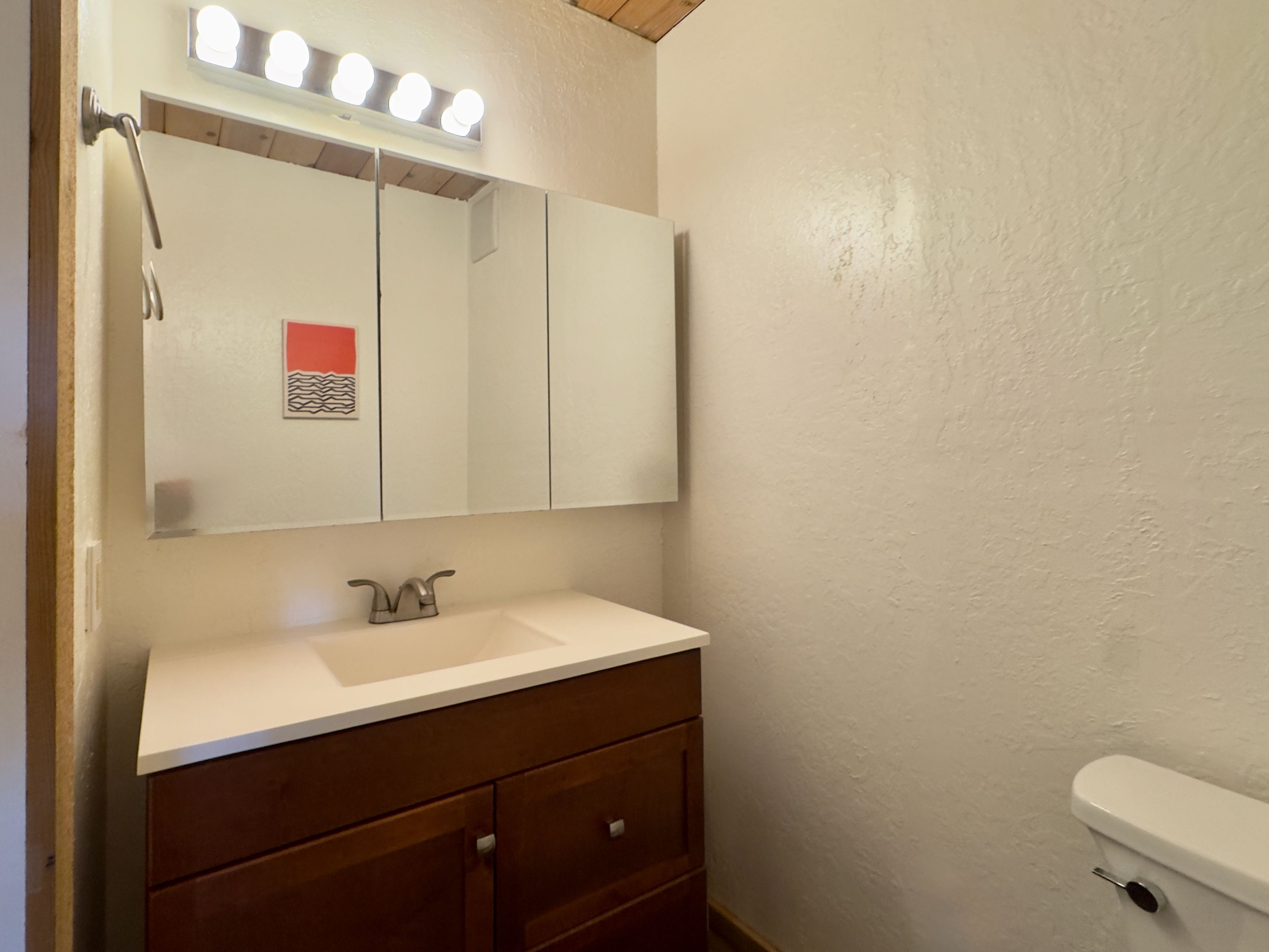 A bathroom with a white sink, a mirror with lights, wooden cabinets, and a toilet.