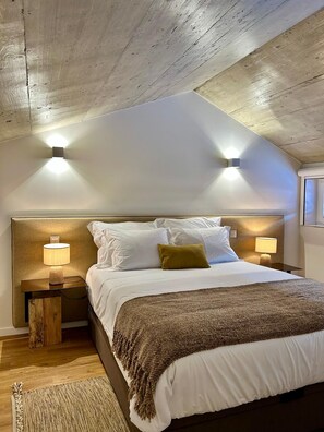 Ein Schlafzimmer mit Holzbalkendecke, einem Bett mit weißer Bettwäsche, zwei Nachttischlampen und einer gewebten Tagesdecke.