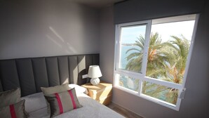A bedroom with a large bed, a nightstand, a lamp, and a window with a view of palm trees and a body of water. - APARTAMENTO VORAMAR by Interhome (Alicante)