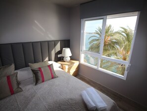Ein Schlafzimmer mit einem großen Bett, einem Nachttisch, einer Lampe und einem Fenster mit Blick auf Palmen und ein Gewässer.