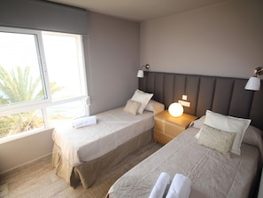 A bedroom with two beds, a nightstand, a bedside lamp, and a window with a view of palm trees. - APARTAMENTO VORAMAR by Interhome (Alicante)