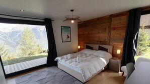 Makuuhuoneessa on suuri sänky, yöpöytä, lamppu, näkymä vuorille ja puinen seinä.