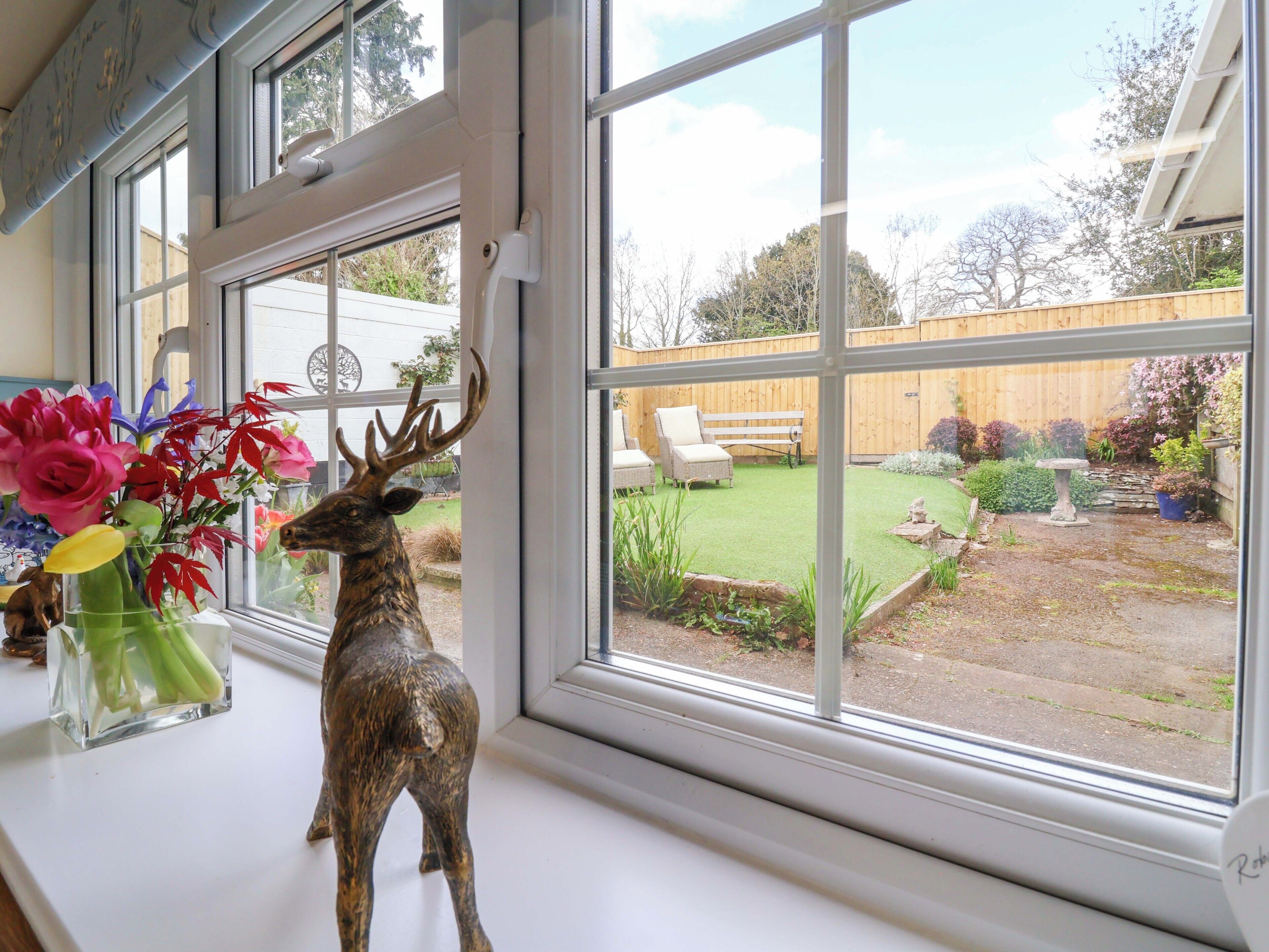 A window with a deer figurine, a vase of flowers, and a view of a backyard with a wooden fence, a bench, and a garden.