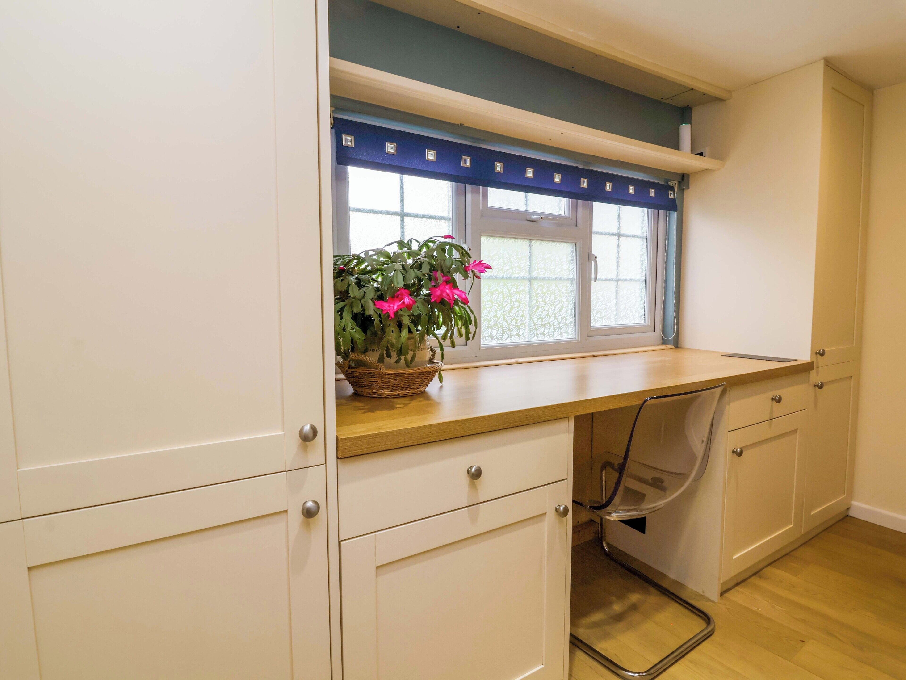 Una cucina con mobili bianchi, un piano di lavoro in legno, una finestra con una tenda azzurra e una pianta in vaso con fiori rosa.