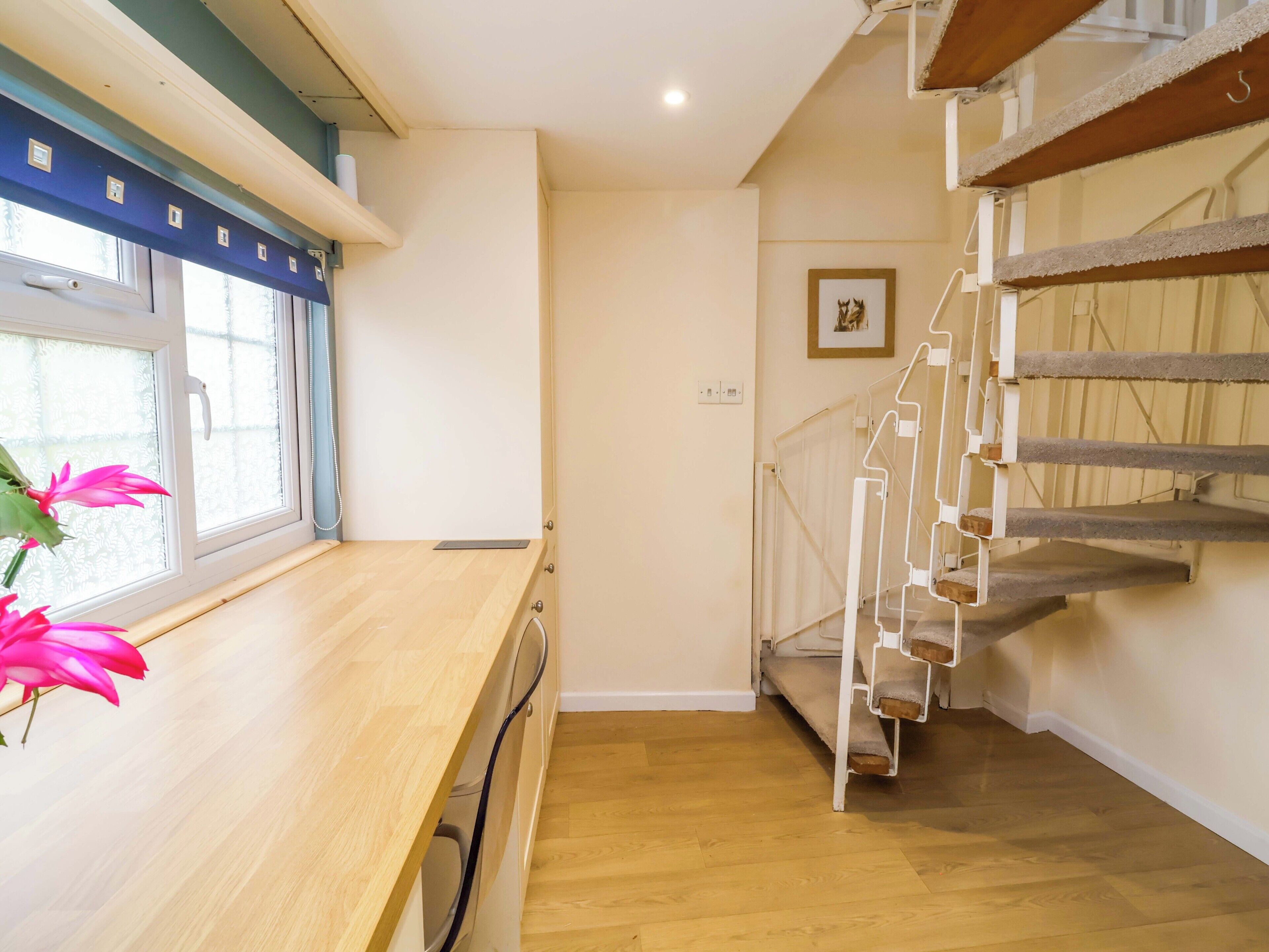 A room with a wooden desk, a window with a view, a staircase, and a framed picture on the wall.
