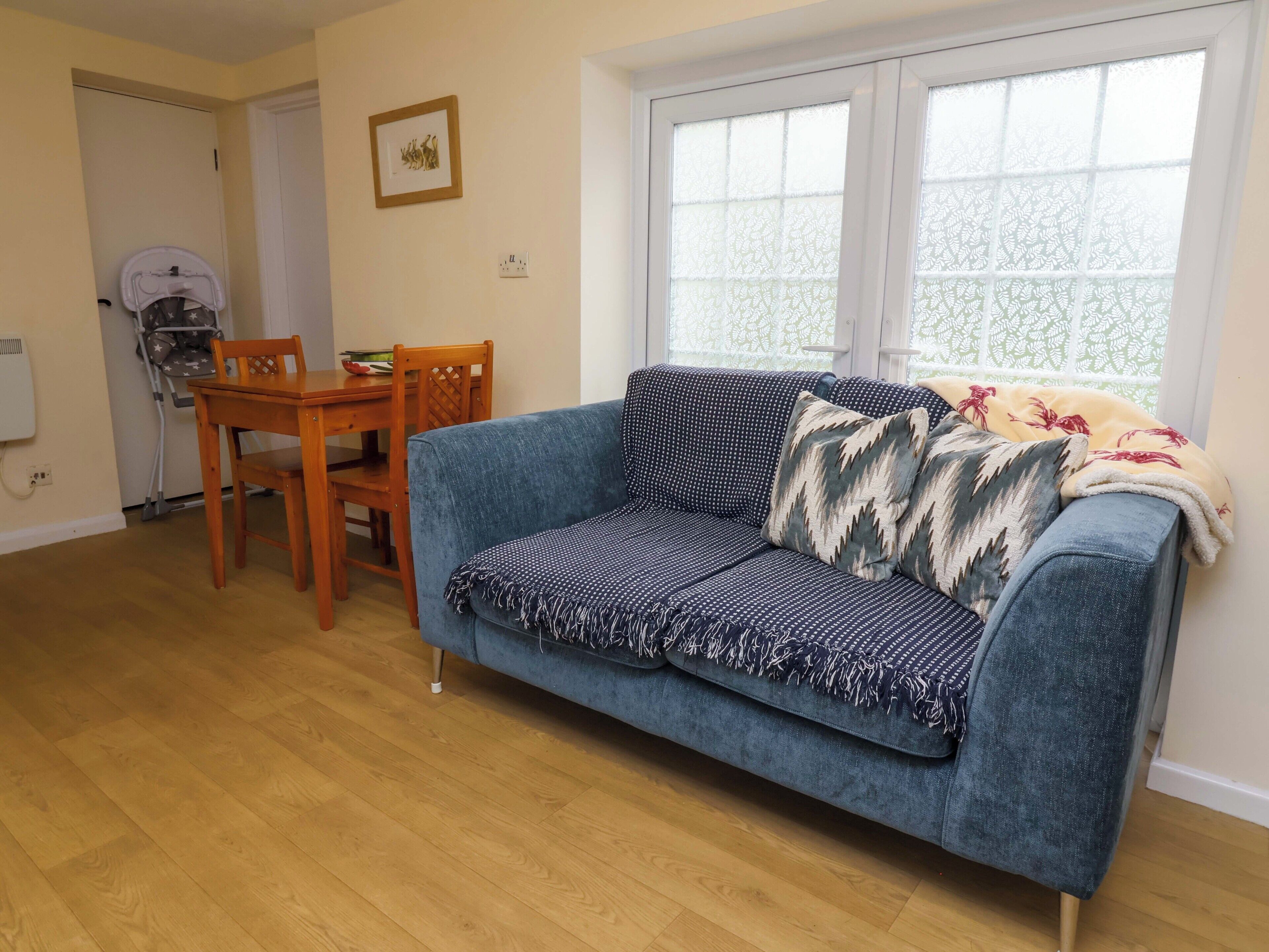A living room with a blue sofa, wooden dining table, chairs, and a window with patterned curtains.