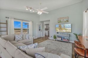 A living room with a sofa, a coffee table, a TV, and a dining area with chairs. - Oceanview | Coastal Haven | Steps to the Beach | 3BR | Fire Pit | 2 10x10 tents & 8 chairs included (Gulf Shores)