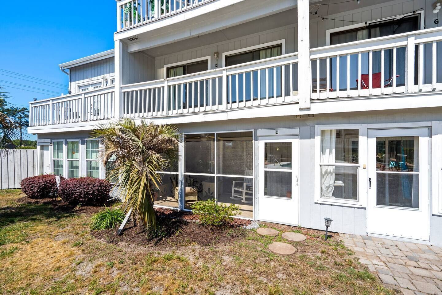 A two-story building with a balcony, a door labeled 'C', and a small garden with a palm tree and shrubs.